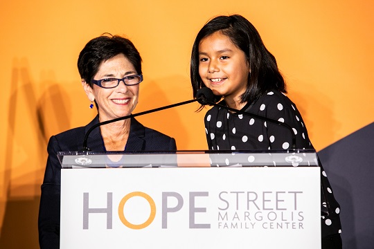 Eloise Appel smiles at a podium as she watches a little girl in polka dots make an announcement